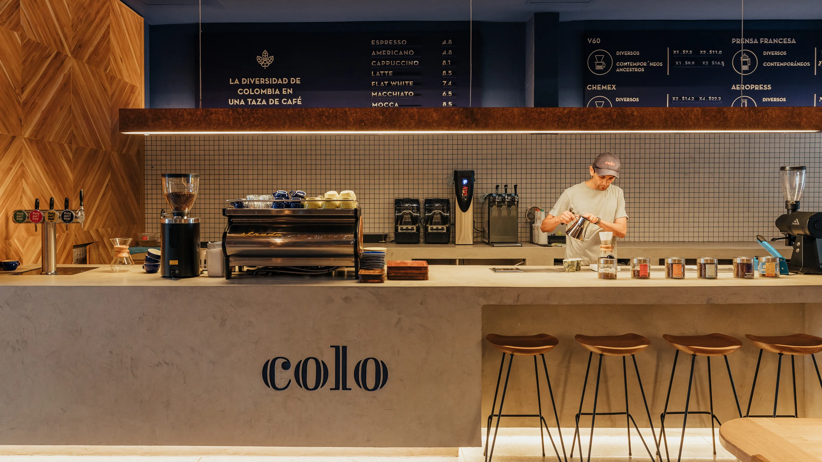 Barra de café con letrero 'colo' junto a un barista preparando una bebida frente a la carta de métodos de preparación y taburetes de madera