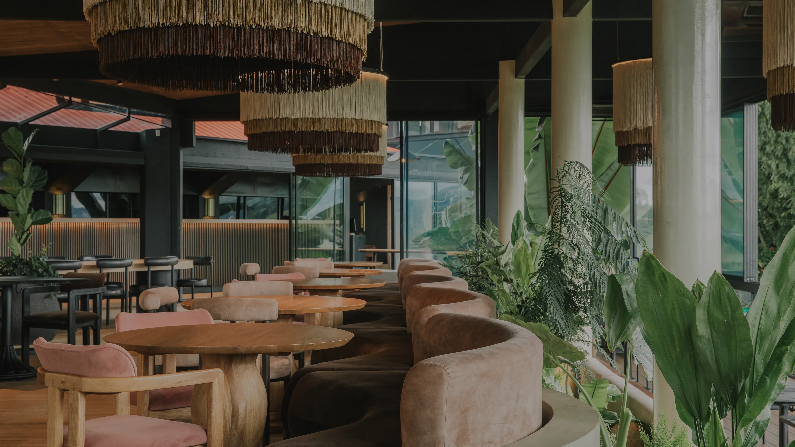 Terraza de restaurante con banca curva tapizada, lámparas de fleco tejidas colgando entre columnas blancas y vegetación tropical
